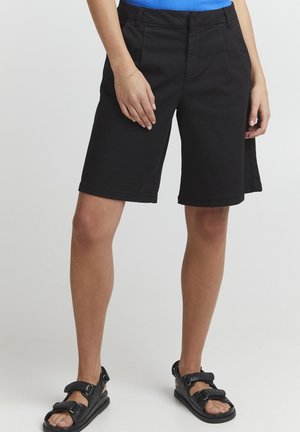 Schwarze Baumwollshorts mit geradem Schnitt, mittellang, mit vorderen Taschen und Gürtelschlaufen. Kombiniert mit schwarzen, schlichten Sandalen mit Riemen.