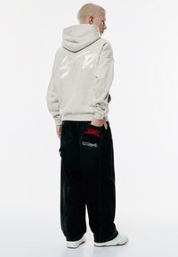Felpa grigia con cappuccio e logo bianco, abbinata a jeans neri a gamba larga con dettagli a fiamma rossa e tasche. Sneakers bianche completano il look.