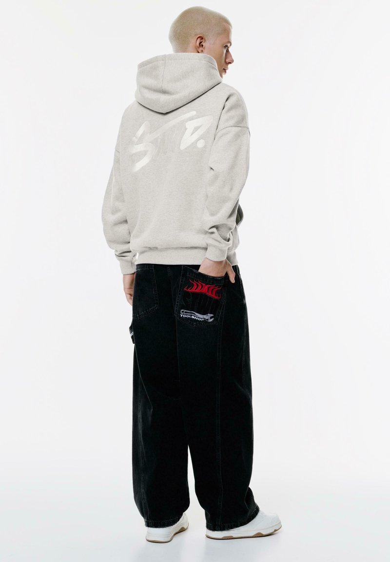 Felpa grigia con cappuccio e logo bianco, abbinata a jeans neri a gamba larga con dettagli a fiamma rossa e tasche. Sneakers bianche completano il look.