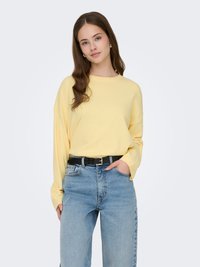 Pull jaune à manches longues en tissu doux, associé à un jean en denim bleu taille haute et une ceinture noire. Coupe droite.