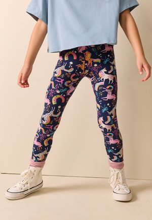Kind trägt marineblaue Leggings mit bunten Einhorn- und Regenbogenmustern, rosa Socken, weiße High-Top-Sneaker und ein hellblaues Oberteil.