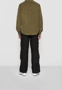 Olijfgroene ribfluwelen shirt met knoopmansetten, gecombineerd met zwarte cargobroeken met zijzakken en een relaxte pasvorm. Witte sneakers maken de look af.