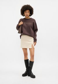 Sweatshirt castanho com o texto "DOLCE FAR NIENTE", saia bege com uma fenda lateral, botas pretas até ao joelho com solas grossas e uma mala castanha.