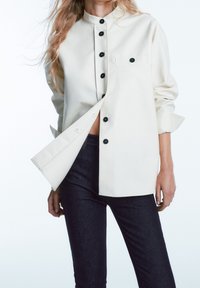 Kremowa koszula na guziki z kołnierzykiem mandarynki, czarnymi guzikami i przednią kieszonką, zestawiona z ciemnymi dżinsami. Prosty i funkcjonalny design.