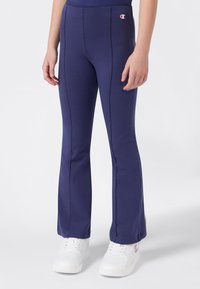 Jongere in marineblauwe getailleerde broek met voorkant naden en witte sneakers met een roze logo aan de zijkant.