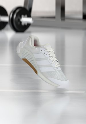 Hvid atletisk sko med mesh og syntetisk overdel, tre sidest stripes og brun sål, der svæver indendørs nær træningsudstyr til vægte.