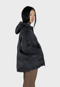 Veste matelassée noire avec capuche, présentant une texture froncée, une fermeture éclair et une coupe surdimensionnée, associée à un pantalon de couleur neutre.