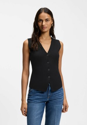 Woman wearing sleeveless black buttoned vest and blue jeans, standing against plain white background.