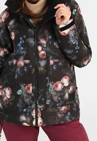 Blommönstrad vinterjacka med svart bakgrund, featuring hög krage, dragkedjestängning och två framfickor. Accentfärger i rosa och grön.