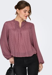 Γυναίκα που φοράει ένα γκοφρέ πουκάμισο σε μωβ αποχρώσεις με λεπτομέρειες δαντέλας, μαύρο παντελόνι, χρυσούς κρίκους στα αυτιά, κρατώντας μια μαύρη τσάντα, κοιτάζει προς τα κάτω.