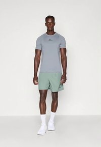 Camiseta deportiva gris con mangas cortas y un logo negro, combinada con pantalones cortos verde claro. El modelo lleva calcetines blancos y zapatillas deportivas.