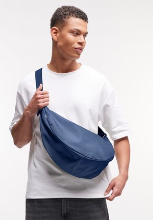 Marineblauer Umhängebeutel mit geschwungener Form und weichem Stoff, verfügt über einen breiten Gurt, dezentes Branding und eine glatte Textur. Für den Freizeitgebrauch entworfen.