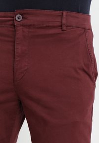 Chino in cotone bordeaux con una vestibilità slim, dotato di chiusura frontale con bottone, tasche laterali e texture liscia.