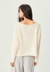 Femme aux longs cheveux bruns portant de grandes boucles d'oreilles, un pull ample couleur écru et un pantalon gris clair, de dos, devant un fond uni.