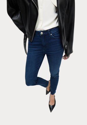 Vrouw die donkeraarblauwe skinny jeans draagt, een wit overhemd met knopen, een zwarte leren jas en zwarte puntige pumps, staand op één been.