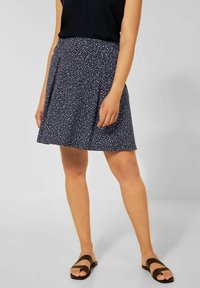 Tmavě modrá sukně s bílými skvrnami, plisovaný design, nad kolena, vyrobená z lehké látky. Kombinována s černým tílkem bez rukávů a hnědými sandály.