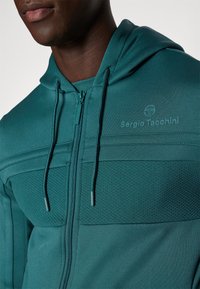 Hoodie zippé en teal avec un panneau texturé, capuche à cordons et logo dans la même couleur. Tissu lisse avec une coupe ajustée.
