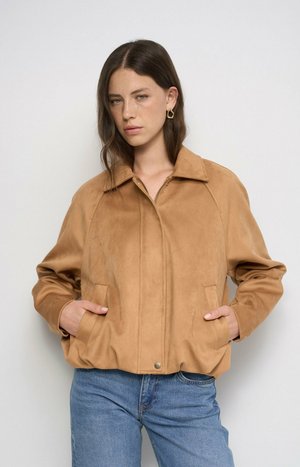 Joven con cabello castaño liso lleva una chaqueta de ante color marrón claro y vaqueros azules, de pie con las manos en los bolsillos de la chaqueta contra un fondo liso.