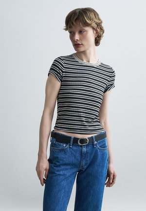 Jeune femme portant un t-shirt à manches courtes rayé noir et blanc, rentré dans un jean bleu avec une ceinture noire, posant devant un fond uni.