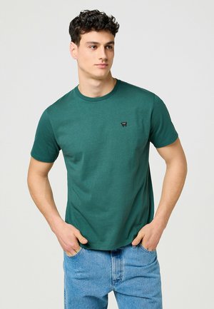 T-shirt vert à manches courtes en tissu texturé, présentant un col rond et un petit logo noir sur la poitrine. Associé à un jean bleu.