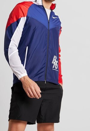 Mann trägt eine farblich abgesetzte Nike-Windjacke in Rot, Weiß und Blau mit Reißverschluss vorne und schwarzen Shorts, steht mit einer Hand in der Tasche.