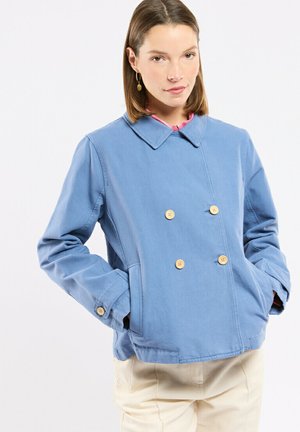 Femme portant une veste bleu clair à double boutonnage avec des boutons en bois, mains dans les poches, et un pantalon de couleur crème sur un fond uni.