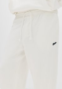Cream-colored sweatpants made of soft fabric with a drawstring waist and a small black logo on the left thigh.