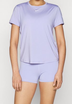 Vrouw die een lichtpaarse Nike-top met korte mouwen en bijpassende strakke shorts draagt, staand tegen een effen witte achtergrond.
