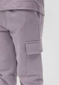 Lila Jogginghosen verfügen über eine weiche Textur, einen elastischen Bund, seitliche Taschen und eine große Cargo-Tasche am Oberschenkel. Einfache Stichdetails sind sichtbar.