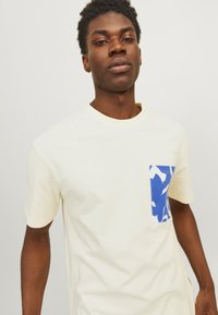 Kortærmet cremefarvet t-shirt lavet af bomuld med en lomme i blå og hvid camouflage mønster på venstre bryst.
