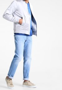 Jasnoszara kurtka bomberka z prążkowanymi mankietami, nałożona na niebieską koszulę. Jasnoniebieskie dżinsy i beżowe sneakersy dopełniają stylizację.