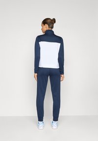 Tamsiai mėlynas ir baltas sportinis treningas, kuriame yra užtrauktuku užsegama striukė su aukštu apykakle ir elastingomis rankovėmis, derinama su atitinkamomis kelnėmis ir sportbačiais.