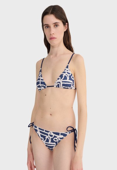 Femme portant un bikini deux pièces à motifs abstraits bleu marine et blanc avec des liens à nouer sur les hanches et les épaules.