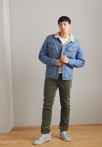 Džinsu jaka ar gaiši zilgu apdari, krēma krāsas kažokādas apkakle, priekšējās kabatas, valkāta pār baltu krekls un kombinēta ar olīvu zaļām biksēm un baltām sporta apaviem.