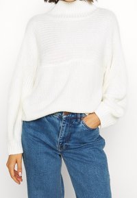 Vit stickad tröja med hög ribbad krage, texturerat mönster och lös passform, tillsammans med blå högmidjade jeans.