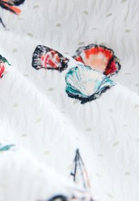 Tela blanca texturizada con patrones de conchas coloridas en naranja, verde azulado y negro, con pequeños puntos decorativos por todo el tejido.