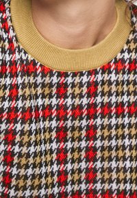 Nahaufnahme eines mehrfarbigen Hahnentrittmuster-Pullovers mit roten, schwarzen, weißen und beigen Formen sowie einem rippierten Kragen in Senfgelb.