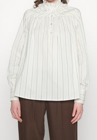 Vit randig blus med volangkrage, knappslå och långa ärmar. Tillverkad av lätt tyg med en avslappnad passform.