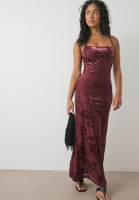 Bordeaux fluwelen maxi-jurk met bloemendessin, dunne bandjes en een aansluitende silhouet, gecombineerd met een zwart handtas met franjes en platte sandalen.