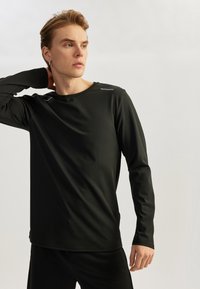 Camisa deportiva de manga larga negra, con textura suave, cuello redondo, diseño ajustado, que presenta un sutil acento reflectante en el hombro.