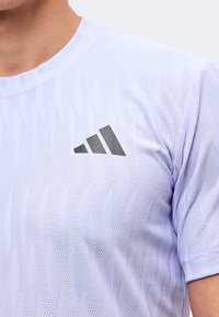 Άτομο που φοράει λευκό αθλητικό μπλουζάκι με ανάγλυφη υφή και μαύρο λογότυπο Adidas στο αριστερό στήθος.