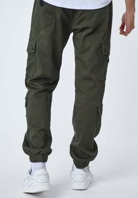 Uomo che indossa pantaloni cargo verde oliva con polsini elastici e sneakers bianche, visto da dietro su uno sfondo neutro.