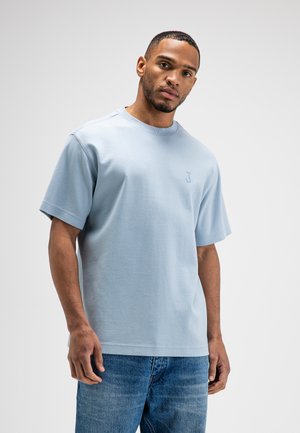 Man die een lichtblauw T-shirt met korte mouwen en een blauwe spijkerbroek draagt, staand tegen een effen lichtgrijze achtergrond.