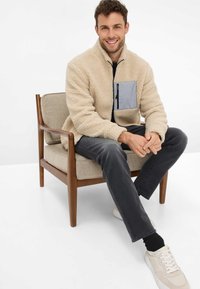 Giacca in pile beige con una tasca con zip grigia, indossata da un uomo seduto su una sedia di legno. Indossa jeans scuri e scarpe da ginnastica chiare.