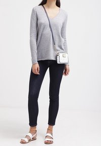 Nainen, joka pukeutuu harmaaseen V-kaula-aukkoiseen villapaitaan, tummiin skinny-farkkuihin, valkoisiin sandaaleihin sekä pieneen valkoiseen olkalaukkuun, jossa on sininen hihna.