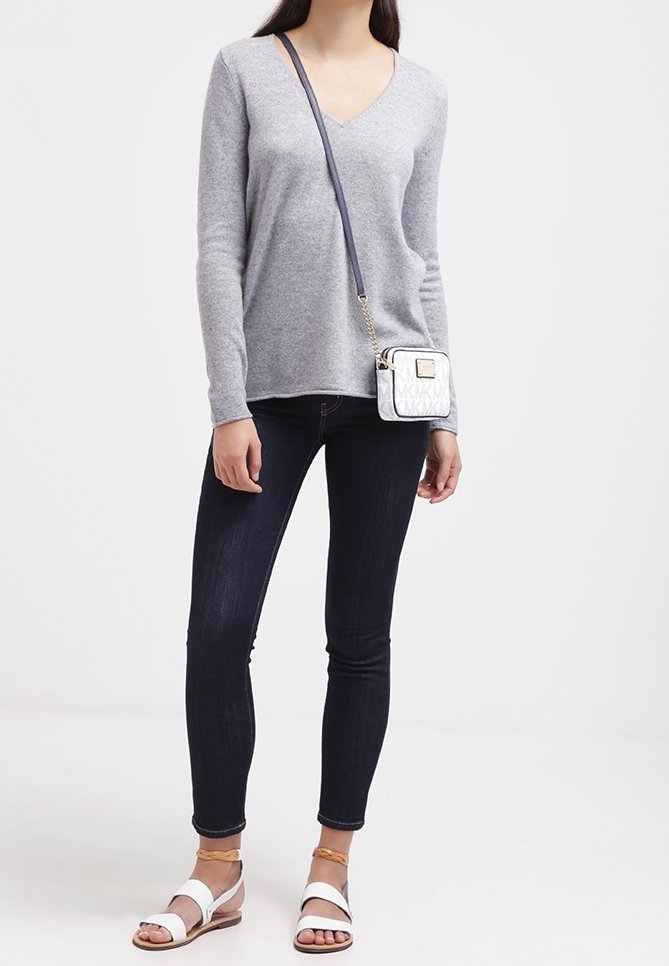 Nainen, joka pukeutuu harmaaseen V-kaula-aukkoiseen villapaitaan, tummiin skinny-farkkuihin, valkoisiin sandaaleihin sekä pieneen valkoiseen olkalaukkuun, jossa on sininen hihna.