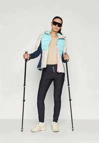 Giacca leggera e colorata con cappuccio, indossata sopra una maglietta beige e pantaloni scuri. Presenta bastoni da trekking e scarpe sportive bianche.