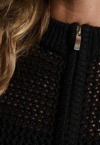 Schwarze gestrickte Zip-Cardigan mit offener Webstruktur, hoher Halslinie und metallic Reißverschluss-Akzent.