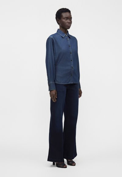 Femme debout portant une chemise en denim bleu foncé boutonnée, un pantalon large bleu marine et des sandales noires à talons sur un fond blanc uni.