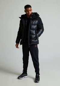 Svart boblejakke med skinnende finish, utstyrt med hette, glidelåslukking og quiltede seksjoner, kombinert med svarte bukser og joggesko.